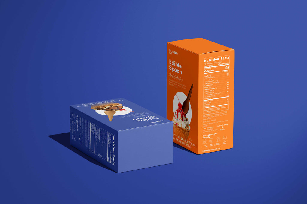 An image of the packaging boxes used by Incredible Eats. There is an orange box (chocolate spoons) standing up and a blue box (vanilla spoons) lying down. It is on a blue background.
