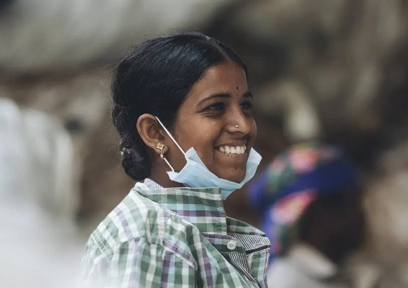 A photo of an Indian woman smiling. She has a mask around her chin and is wearing a green and white plaid shirt.